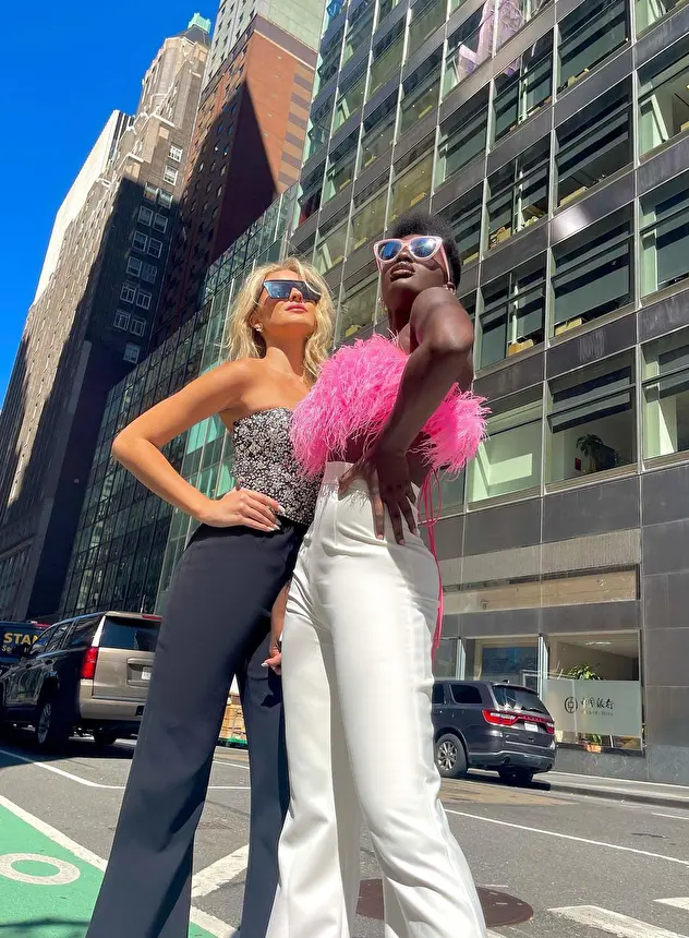 Two models on a sunny street: one blonde in a Jovani black and white embellished top and trousers, the other in a pink Jovani top (Style 09835) and white trousers. 