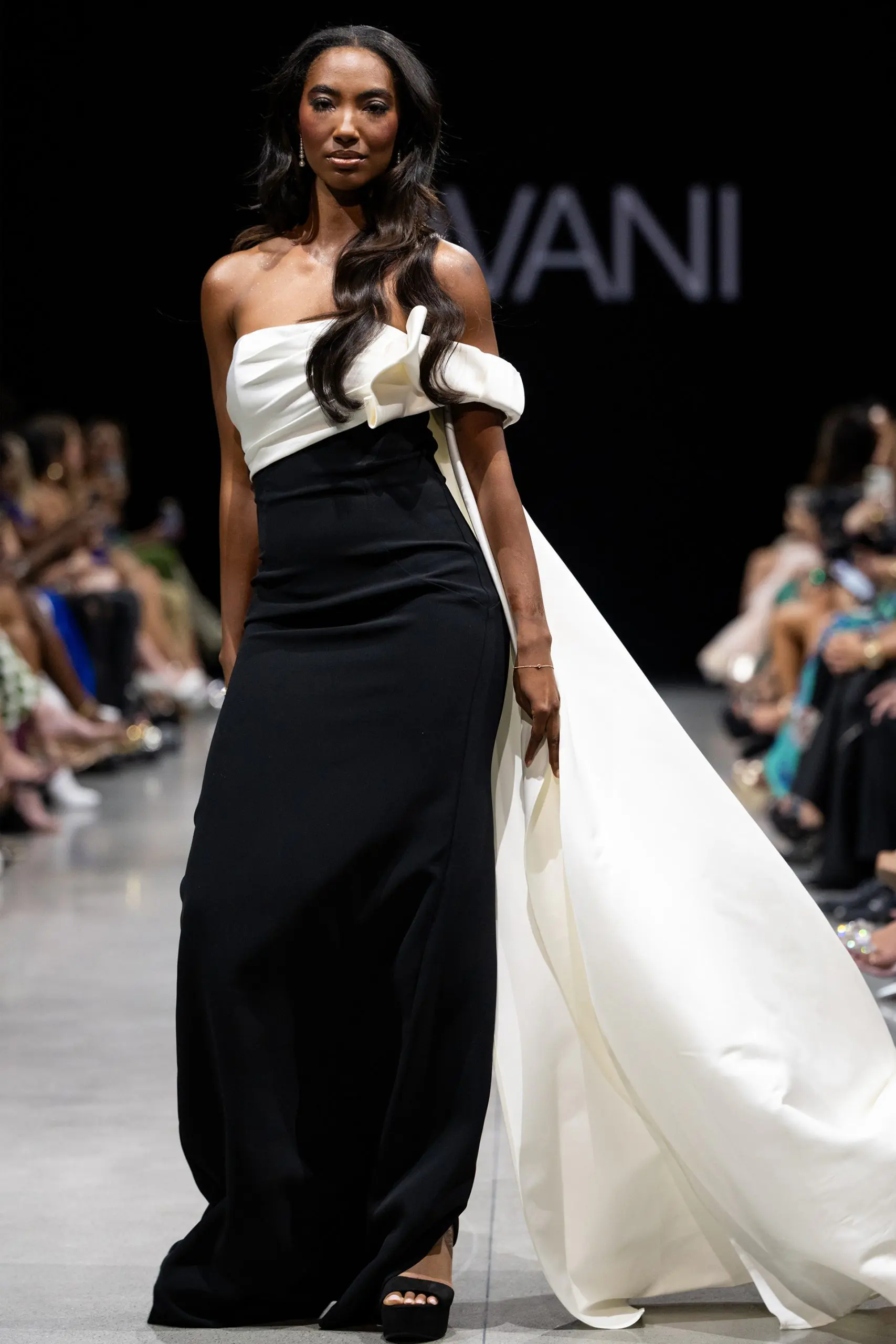 Model wearing Jovani AT38864 elegant black gown with asymmetrical neckline and cape-like detail.