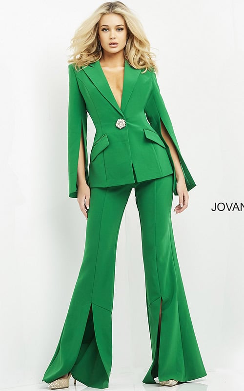 Model wearing Jovani 06922 emerald green jumpsuit with cape sleeves and brooch detail, front view.