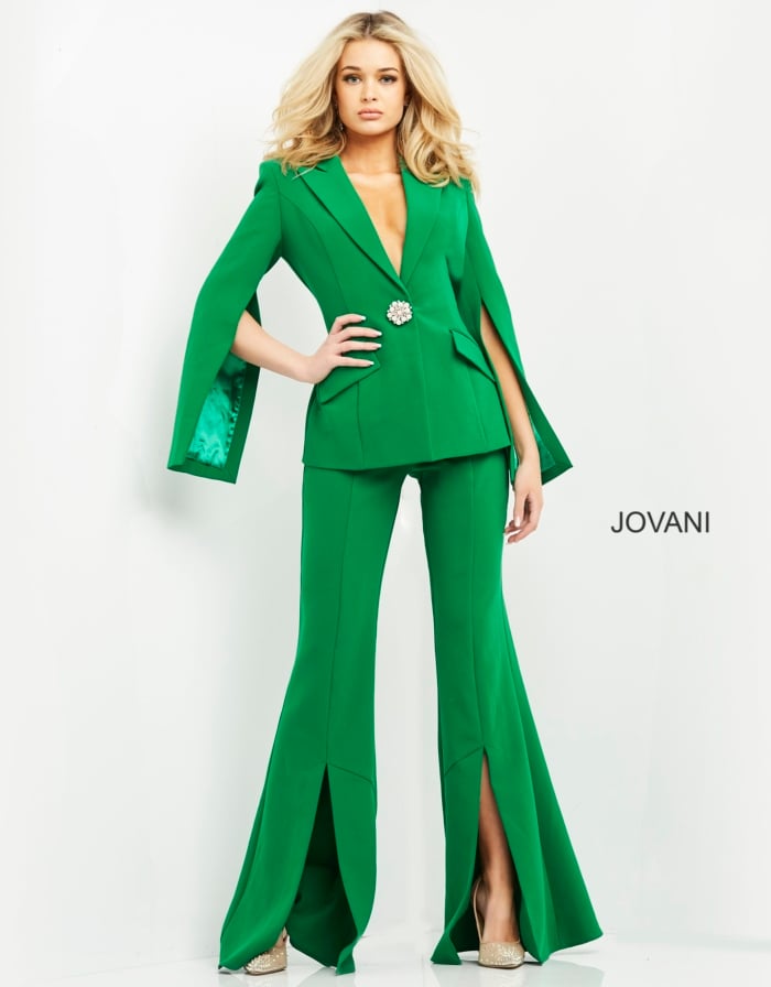 Model wearing Jovani 06922 emerald green jumpsuit with cape sleeves and brooch detail, front view.