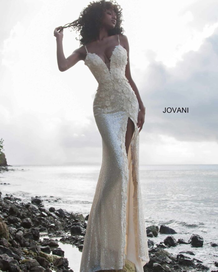 Model wearing Jovani 1012 white gown with floral details and a high slit by the seaside.