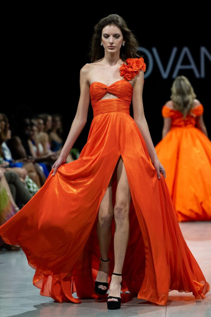 Model wearing Jovani 37254 orange gown with one-shoulder floral applique and high slit.