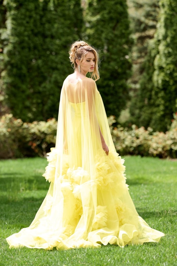 Model wearing Jovani 40092 yellow gown with flowing tulle and floral details, angled back view