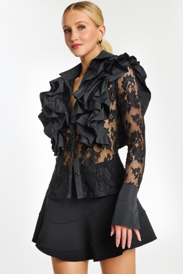Model in black floral lace top with exaggerated ruffle shoulders and structured black skirt