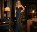 Model wearing a fitted, dark green shimmering evening gown with ruched fabric detail in a dimly lit lounge.