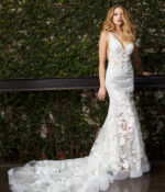 Model in Jovani JB04192 white gown with floral design standing next to greenery