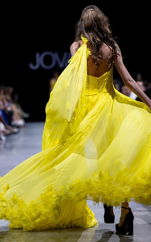 Model wearing Jovani S38104 yellow gown with open back and dramatic cape, back view.