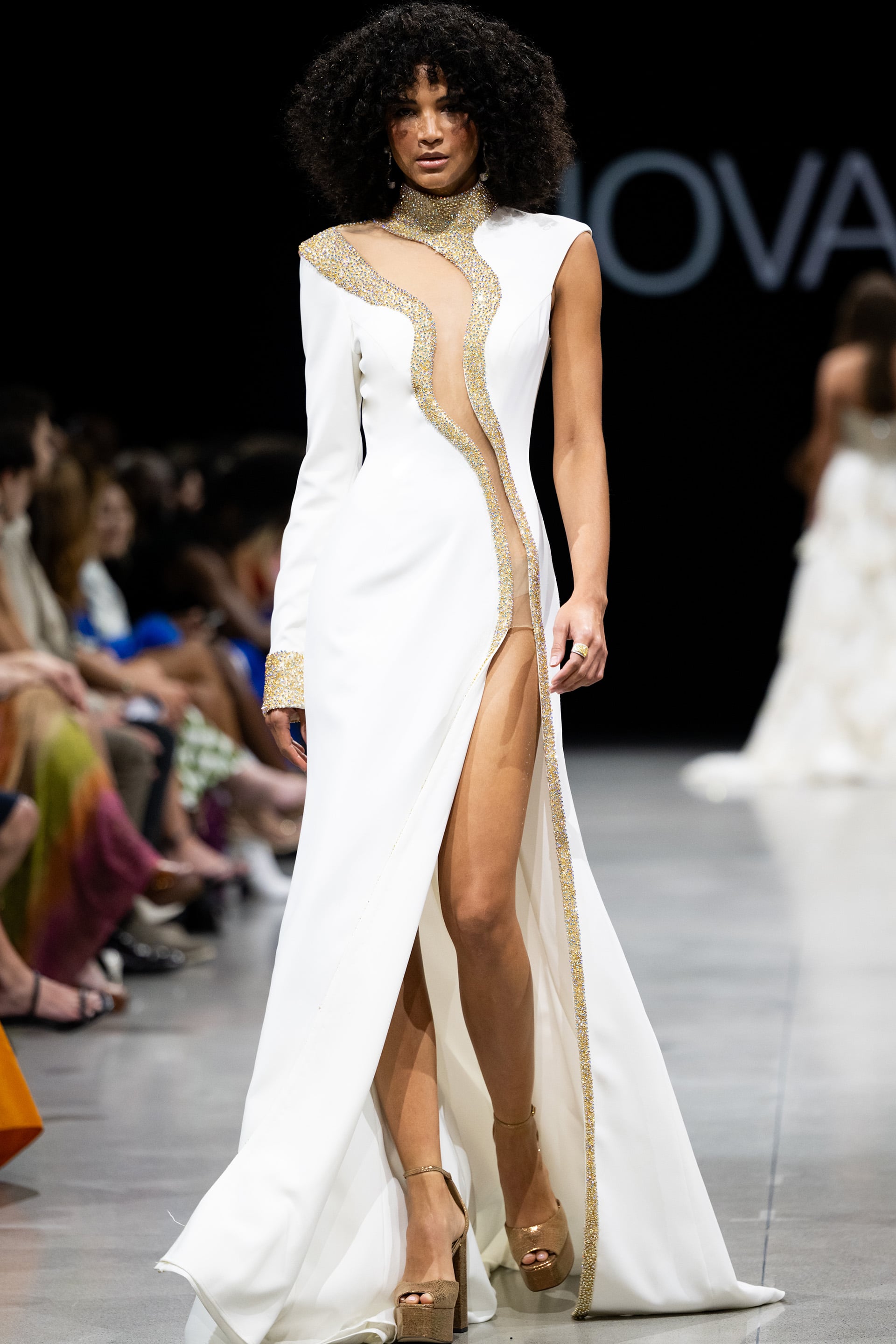 S38446.jpg Model wearing Jovani S38446 white gown featuring a high neckline, embellishments, high slit, and full-length sleek design.